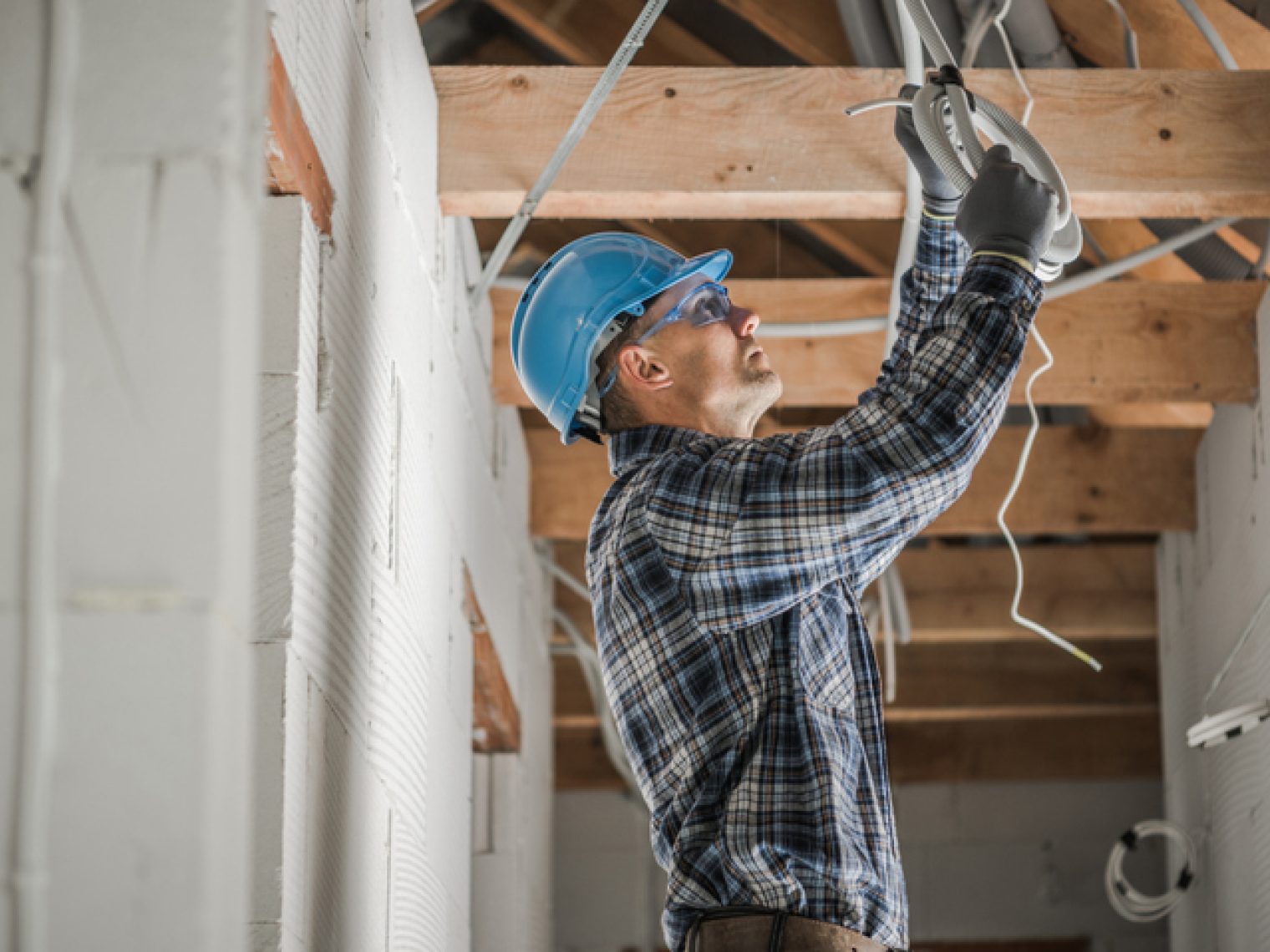 A focused construction worker wearing a hard hat and protective gloves is installing or repairing wiring or fixtures in a house under construction or renovation.