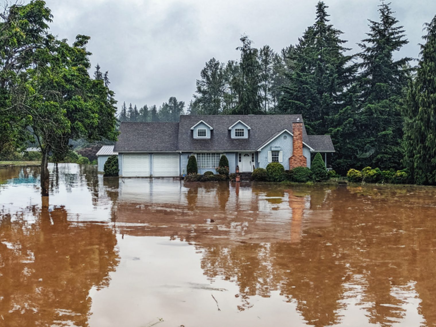 A suburban home is surrounded by floodwaters, with trees reflecting in the still water, under a gloomy sky, highlighting the impact of severe weather.A suburban home is surrounded by floodwaters, with trees reflecting in the still water, under a gloomy sky, highlighting the impact of severe weather.