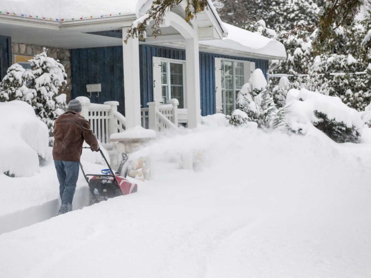 A person in winter clothing uses a snowblower to clear a thick layer of snow in front of a house with snow-covered trees and landscape.