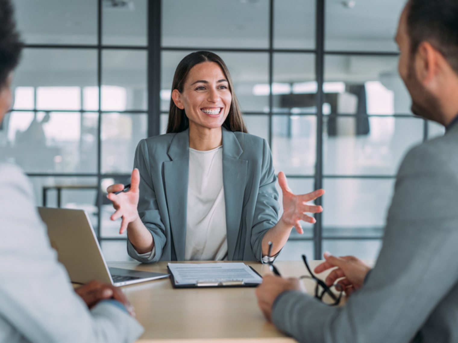 A cheerful professional woman speaks with enthusiasm during a meeting with two other individuals in a bright office with a cityscape visible through the windows behind her.